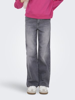 Persona que lleva vaqueros anchos gris descoloridos, zapatillas blancas y una sudadera rosa brillante, de pie contra un fondo claro y liso.