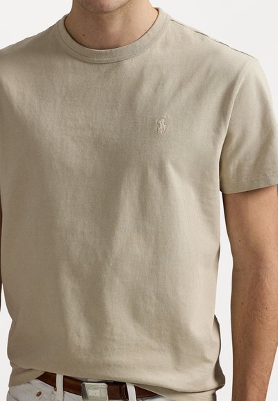 Beige bomullst-shirt med rund hals, korta ärmar och en liten broderad logotyp på vänster bröst. Slät textur, avslappnad passform.