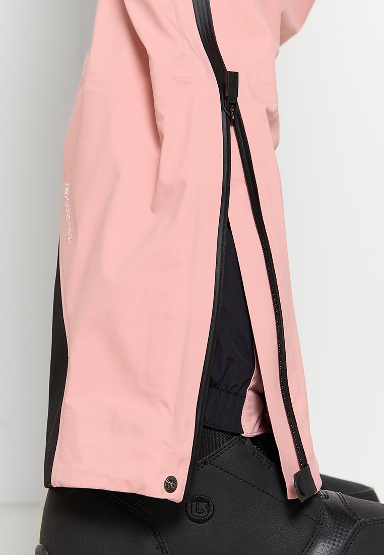 Rosa wasserdichte Jacke mit einem schwarzen Reißverschluss, elastischen Bündchen und glatter Oberfläche, getragen über einem schwarzen Stiefel.