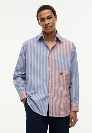 Junger Mann trägt ein gestreiftes Hemd mit geteilten Farben in Blau und Pink, mit Fronttasche und dunkler Hose, posiert vor weißem Hintergrund.