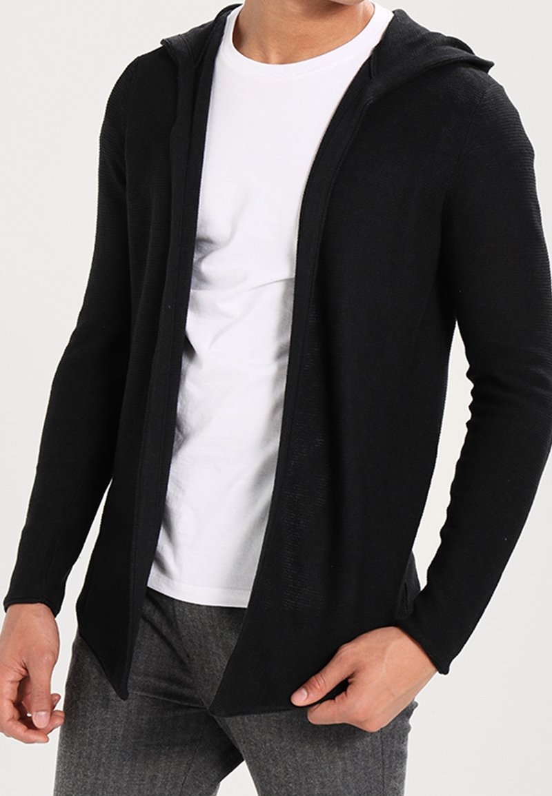 Man som bär en öppen svart huva cardigan över en vit t-shirt och grå byxor, står med handen som håller i kantens på cardigan.