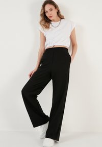 Schwarze weite Hose mit elastischem Bund, kombiniert mit einem weißen cropped T-Shirt. Das T-Shirt hat geraffte Details am Saum.
