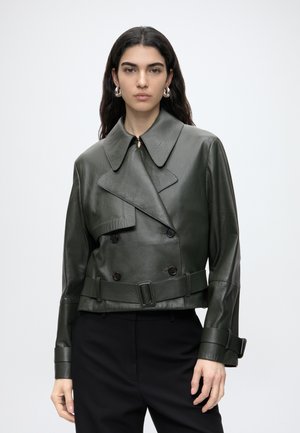 Femme aux longs cheveux foncés porte une veste en cuir vert foncé à double boutonnage avec large col et ceinture, associée à un pantalon noir et des boucles d'oreilles argentées en anneau.