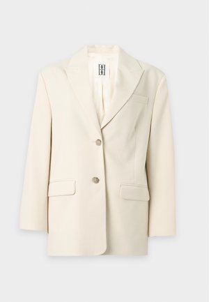 Vrouw met kort haar draagt een crèmekleurige blazer, wit T-shirt, beige hoge taille wijde broek en bruine hoge hakken, staat met handen in de zakken.