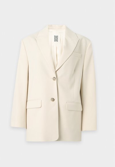 Blazer sur mesure de couleur crème avec revers crantés, deux boutons et poches à rabat devant sur un fond uni.