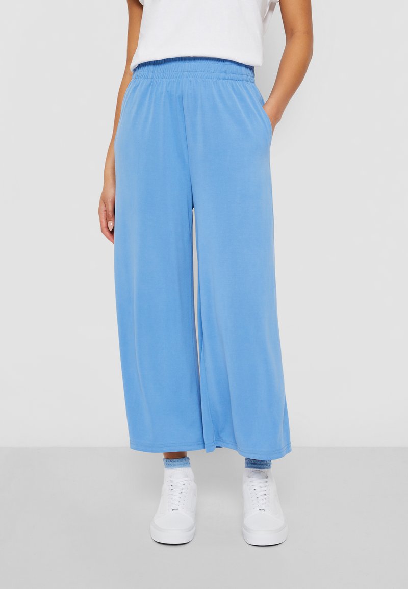 Pantalones de pierna ancha en azul claro con cintura elástica, dos bolsillos laterales y una textura suave, combinados con zapatillas blancas.