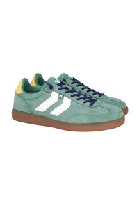 Zapatillas de ante verde con rayas blancas, cordones azul marino y acentos amarillos en el talón. Presenta una suela de goma texturizada marrón.