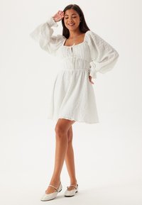Vit, texturerad klänning med fyrkantig halsringning, puffärmar och resår i midjan. Kombinerad med ljusa, platta skor.
