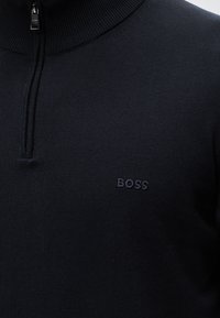 Czarny sweter z zamkiem do ćwiartki, prążkowanym kołnierzem i małym, wyhaftowanym logo "BOSS" na lewej piersi.