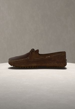 Brown suede leather loafer with moc toe stitching, side laces, and rubber-studded sole displayed on a neutral surface and background.