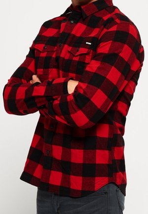 Mann trägt ein langärmeliges Hemd mit rotem und schwarzem kariertem Muster und Brusttaschen, die Arme verschränkt, und dunkle graue Jeans.