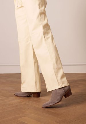 Stivaletti alla caviglia grigi in suede con punta affusolata e tacco in legno impilato, abbinati a pantaloni color crema su un pavimento in legno.