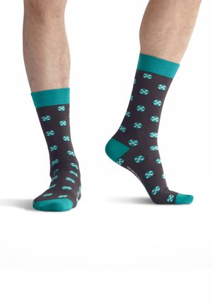 Paar Beine mit dunkelgrauen Socken mit türkisfarbenen Bündchen, Fersen und Zehen, mit einem wiederholenden türkisfarbenen und weißen geometrischen Muster.