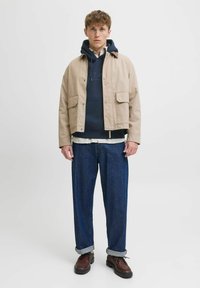 Chaqueta beige claro con dos bolsillos, superpuesta a una sudadera azul marino. Pantalones de mezclilla azul oscuro con puños enrollados y zapatos marrones con cordones.