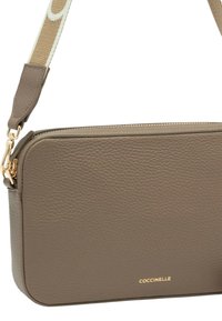 Taupefarbene Umhängetasche aus strukturiertem Leder mit goldfarbenem Reißverschluss, goldfarbenen Beschlägen, beigem, verstellbarem Stoffriemen und COCCINELLE-Logo in goldener Schrift.