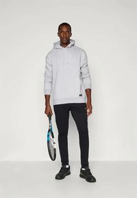 Γκρι φούτερ με καγκουρό τσέπη και κορδόνι, σε συνδυασμό με μαύρο αθλητικό παντελόνι. Το μοντέλο κρατάει μια ρακέτα του τένις, φορώντας μαύρα αθλητικά παπούτσια.