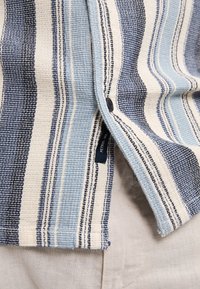 Gestreept shirt van geweven stof in blauw en crèmekleuren, met knopen en een label, dat een textuur met gevarieerde patronen toont.