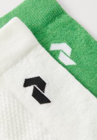 Dva para sportskih čarapa: jedan zeleni s bijelim logom i jedan bijeli s crnim logom, s teksturiranim materijalom i rebrastim rubovima.