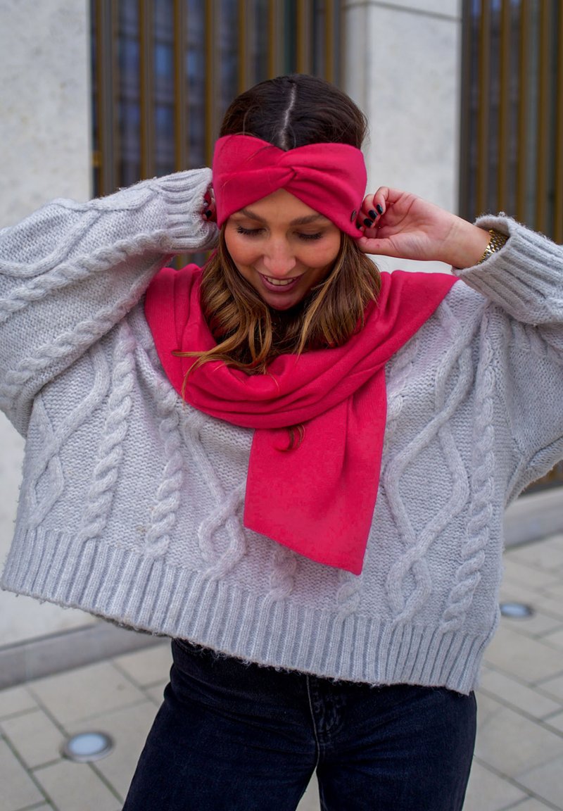 Pull en maille torsadée gris clair avec une coupe oversized, associé à une écharpe rose vibrante et un serre-tête torsadé. Le modèle ajuste les accessoires.
