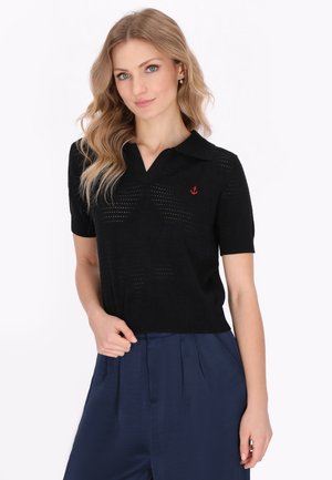 Černý polo svetr s krátkým rukávem s texturovaným vzorem a červeným logem kotvy na hrudi. Stylizováno s širokými námořnickými kalhotami.