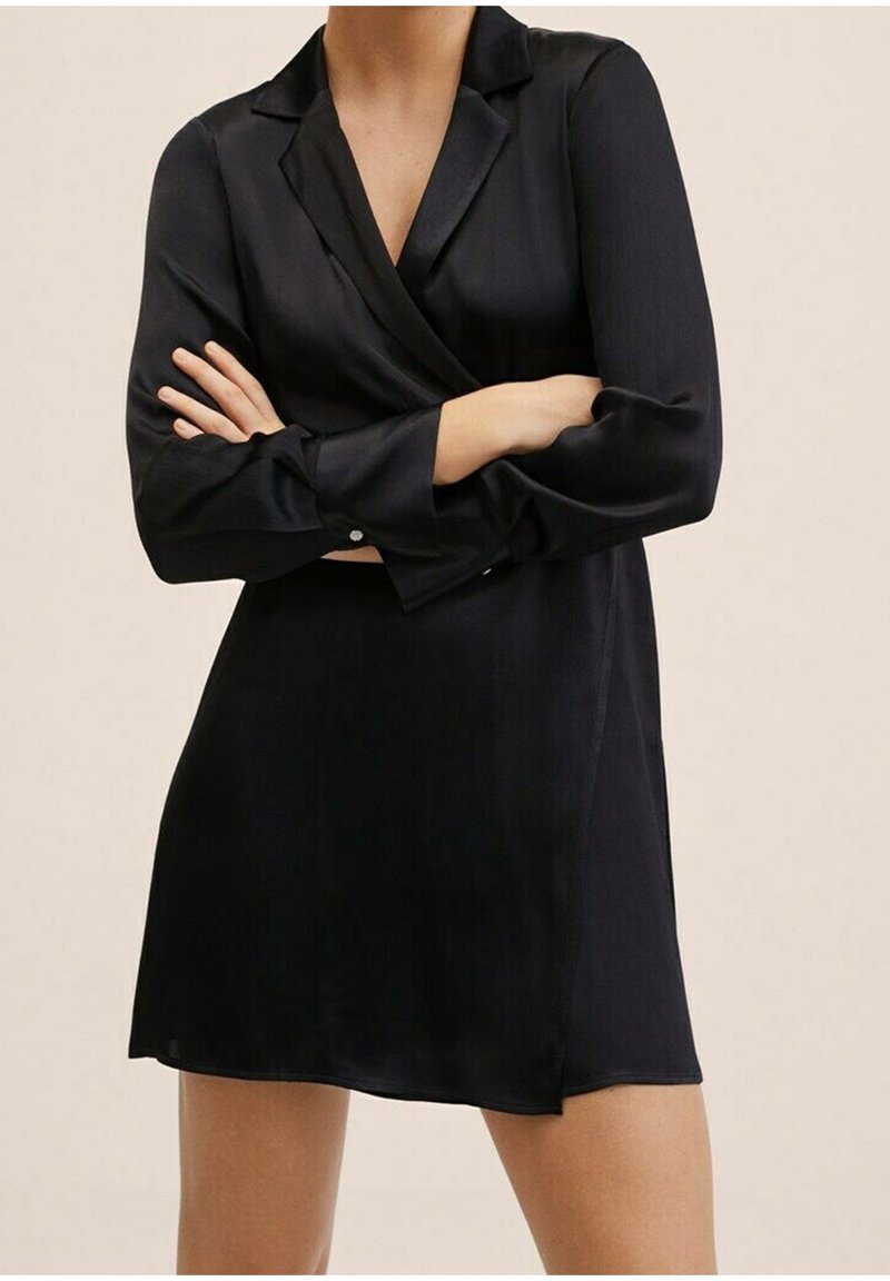 Femme portant une robe portefeuille en satin noir à manches longues avec col, bras croisés, cadrée au niveau du cou et à mi-cuisse.