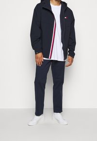 Navy windbreaker jacket with a hood, featuring a front zipper with red and white stripes, paired with a white T-shirt and navy pants.