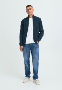 Marineblå zip-genser over hvit t-skjorte, kombinert med blå jeans og hvite sneakers. Strikketeksturen er synlig på genseren.