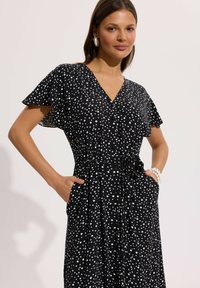 Kobieta w czarnej, krótkorękawowej sukience w białe kropki, z przodu na guziki, wiązana w talii, z rękami w kieszeniach, nosząca perłową bransoletkę i duże kolczyki.