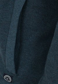 Mörkblå stickad tröja med en strukturerad yta och en etikett med en cirkulär logotyp. Har en rund halsringning och långa ärmar.