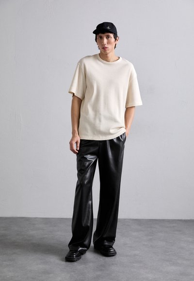 Beige strukturierte Kurzarmbluse kombiniert mit schwarzen weiten Hosen aus Kunstleder und schwarzen klobigen Schuhen, vor einem neutralen Hintergrund.