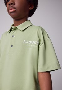 Lysegrøn poloshirt lavet af blødt stof. Har en klassisk krave, tre knapper og et trykt logo på brystet i hvid.