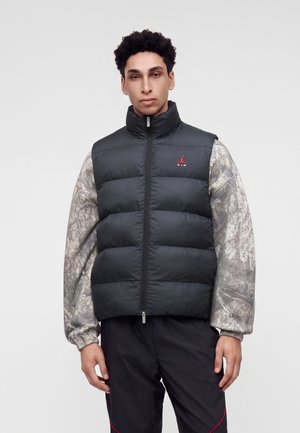 Joven con chaleco acolchado negro sobre una camisa gris de manga larga con estampado y pantalones negros, de pie frente a un fondo liso.