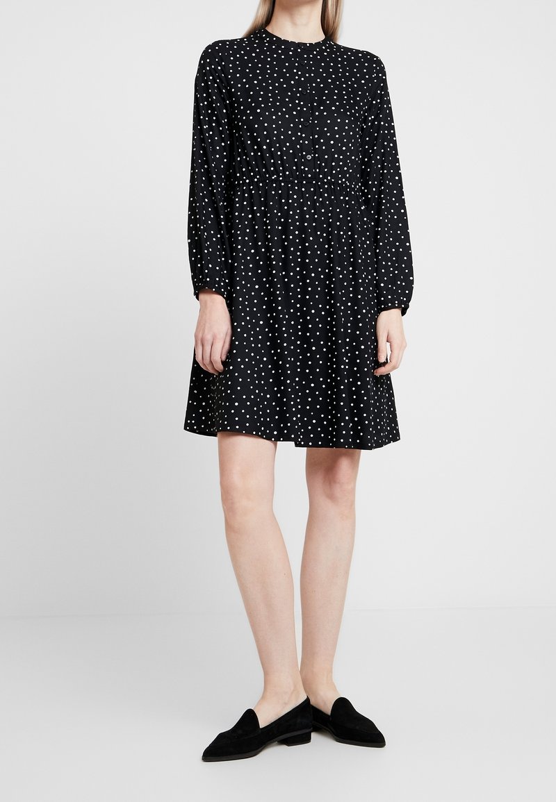 Robe noire à pois blancs, manches longues, col rond et taille froncée. Associée à des chaussures à enfiler ensuède noire.