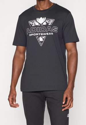 Mann trägt schwarze Adidas Sportbekleidung mit einem floralen Dreieckslogo auf dem T-Shirt und passenden schwarzen Hosen.