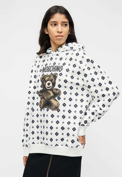 Λευκή φούτερ με κουκούλα με γραφικό αρκουδάκι με μαύρες και χρυσές λεπτομέρειες, περιβαλλόμενο από μοτίβο μπλε διαμαντιών. Απαλό ύφασμα με άνετη εφαρμογή.