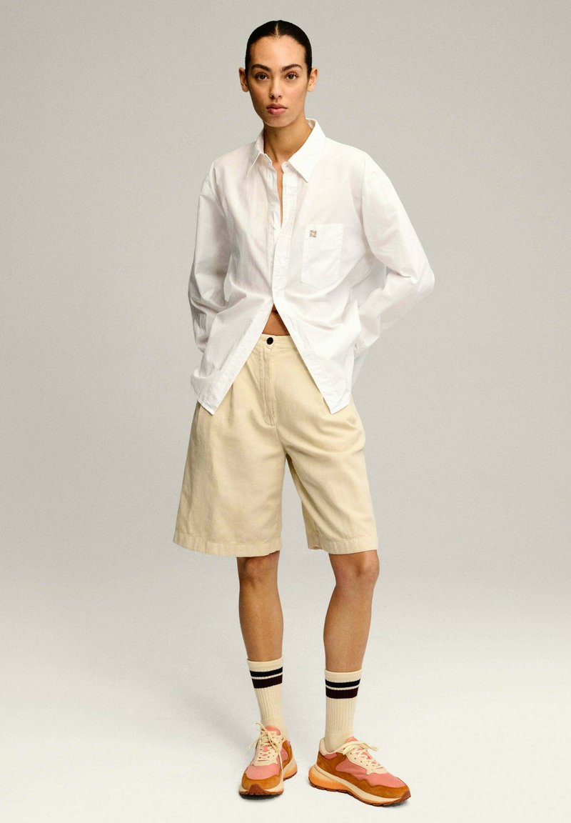 Camisa blanca de botones, pantalones cortos beige y zapatillas multicolores. Las características incluyen un bolsillo en la camisa, calcetines a rayas y un corte relajado.