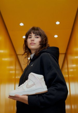 Młoda osoba w czarnej bluzie z kapturem trzyma białego sneakersa za kostkę z beżowymi akcentami na tle żółtej ściany z oświetleniem sufitowym.
