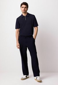 Polo shirt blu navy con colletto a zip, tessuto strutturato, abbinato a pantaloni scuri e sneakers bianche con accenti grigi e neri.