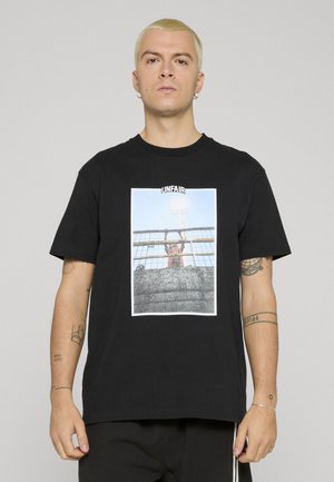 Mann med kort blondt hår iført svart T-skjorte med bilde av person bak gjerde med teksten «URETTFERDIG» over.