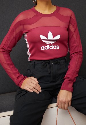 Kobieta w bordowej, siateczkowej bluzce z długim rękawem marki Adidas z białym logo oraz czarnych, wysokich spodni, siedzi z jedną ręką na kolanie.
