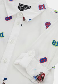 Witte overhemd met knopen, voorzien van kleurrijke borduursels, waaronder cijfers en een grafisch detail. De stof is glad met een gestructureerde kraag.