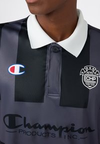 Zbliżenie na czarno-szaro paskowaną koszulkę polo z białym kołnierzykiem, logo Champion na lewej piersi i emblemat NYC CHP 1919 na prawej piersi.