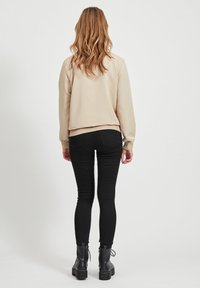 Frau mit langen, gewellten Haaren steht mit dem Rücken zur Kamera, trägt einen beigen Sweatshirt, schwarze Skinny-Jeans und schwarze Schnürstiefel vor einem einfarbigen Hintergrund.