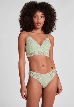 Frau mit lockigem Haar, die ein hellgrünes Spitzen-Bralette und passende Spitzenunterhosen trägt, vor einem schlichten Hintergrund stehend.