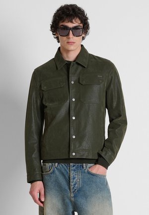 Jonge man met een donkergroene jasje met knopen, blauwe vervaagde jeans en grote zwarte zonnebril, staand tegen een effen lichte achtergrond.