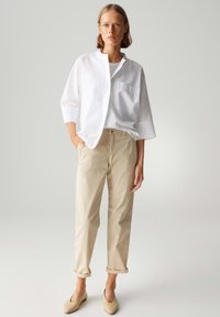 Weißes, kragenloses Hemd mit einer Fronttasche, kombiniert mit hochgekrempelten beige Hosen und hellbeigen Slippern. Glatte Stofftexturen.