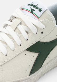 White leather sneaker with green accent logo, textured surface, padded tongue, and white laces. Notable side perforations for ventilation.