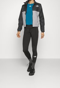 Chaqueta negra y gris con cremallera, top color aguamarina, leggings negros con logo y zapatillas oscuras, creando un atuendo deportivo en capas con texturas variadas.