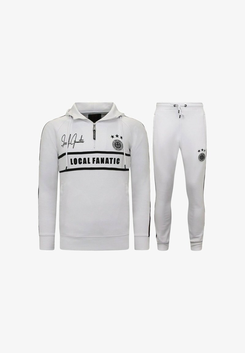 Wit trainingspak bestaande uit een pullover met een ritssluiting bij de hals en een broek. Voorzien van zwarte strepen, logo details en drie sterren op het bovenstuk.