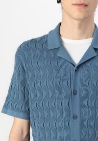 Short-sleeve teal polo shirt made of knitted fabric featuring a wave pattern, with a collar and black buttons.